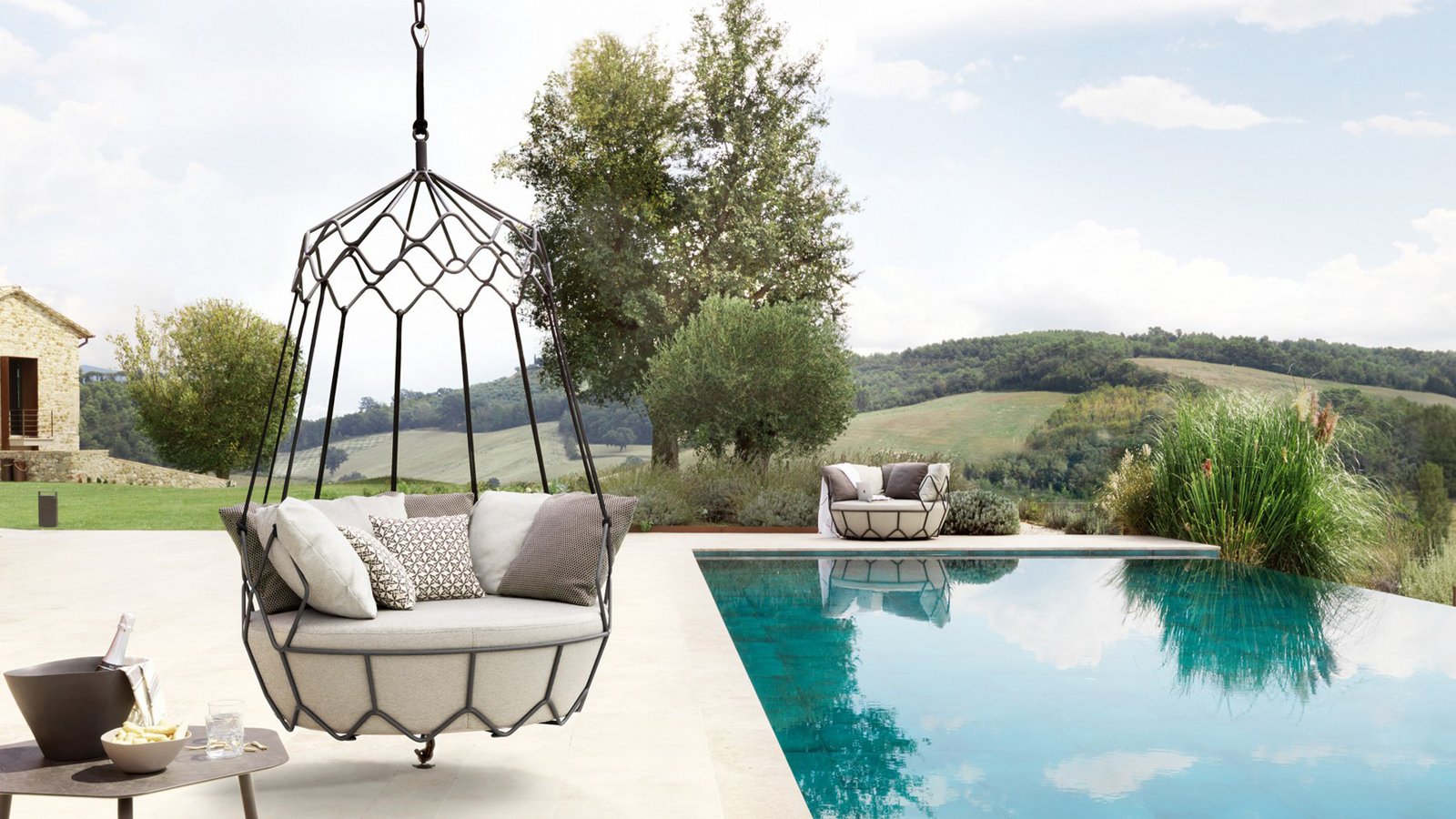 Modern outdoor hanging daybed with plush cushions on a poolside patio overlooking a scenic hillside landscape.