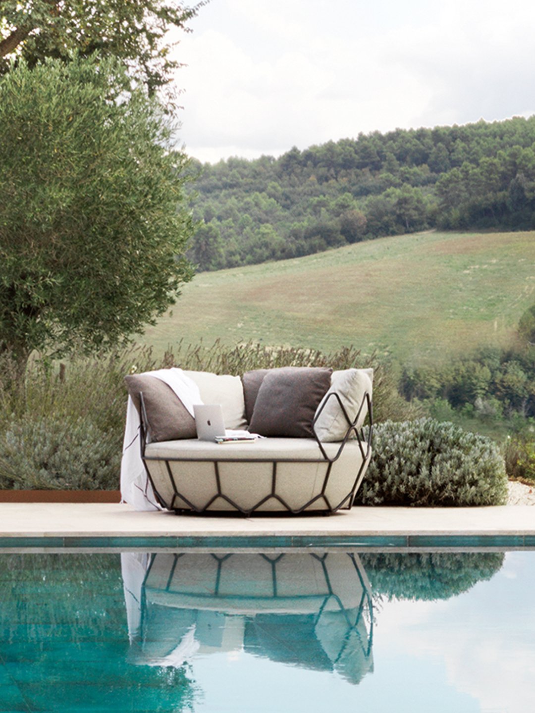 Modern circular outdoor daybed with geometric metal frame poolside, reflecting in clear blue water.
