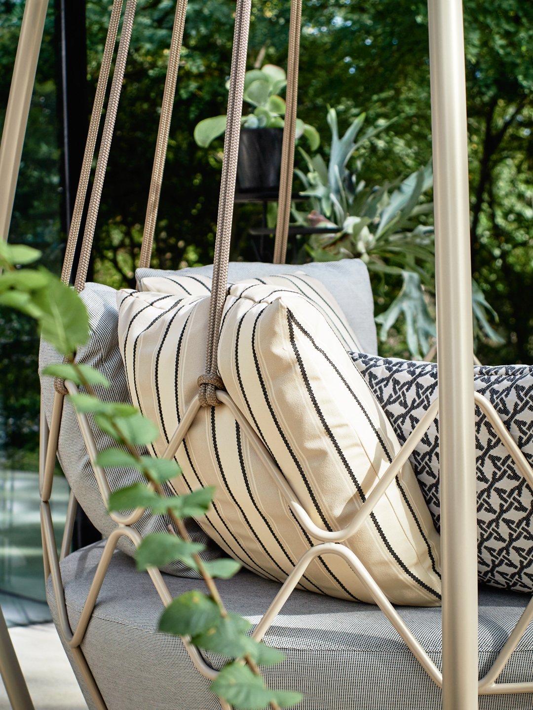 Close-up of a designer outdoor swing chair featuring elegant striped cushions and a beige metal frame in a lush garden.